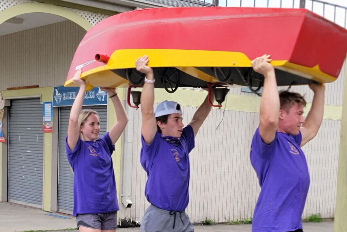 Two male students 和 a female student carry a boat above their heads across Eldridge Park during a community service outing